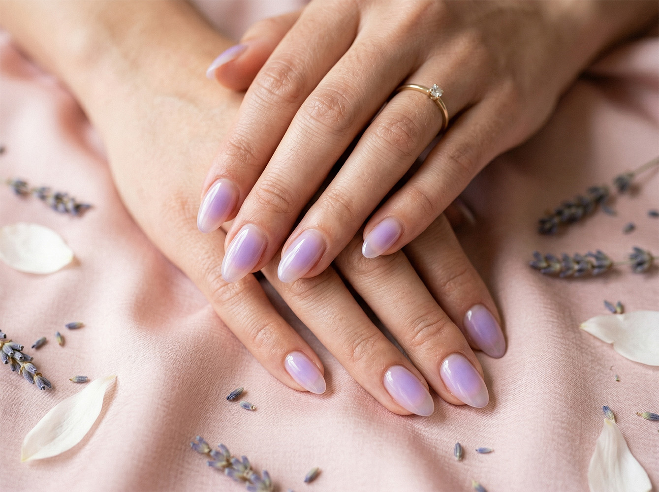 Zbliżenie na paznokcie w modnym stylu aura nails z delikatnym lawendowym środkiem rozmywającym się do przezroczystych brzegów. Dłonie ułożone na jasnoróżowym jedwabiu z gałązkami lawendy