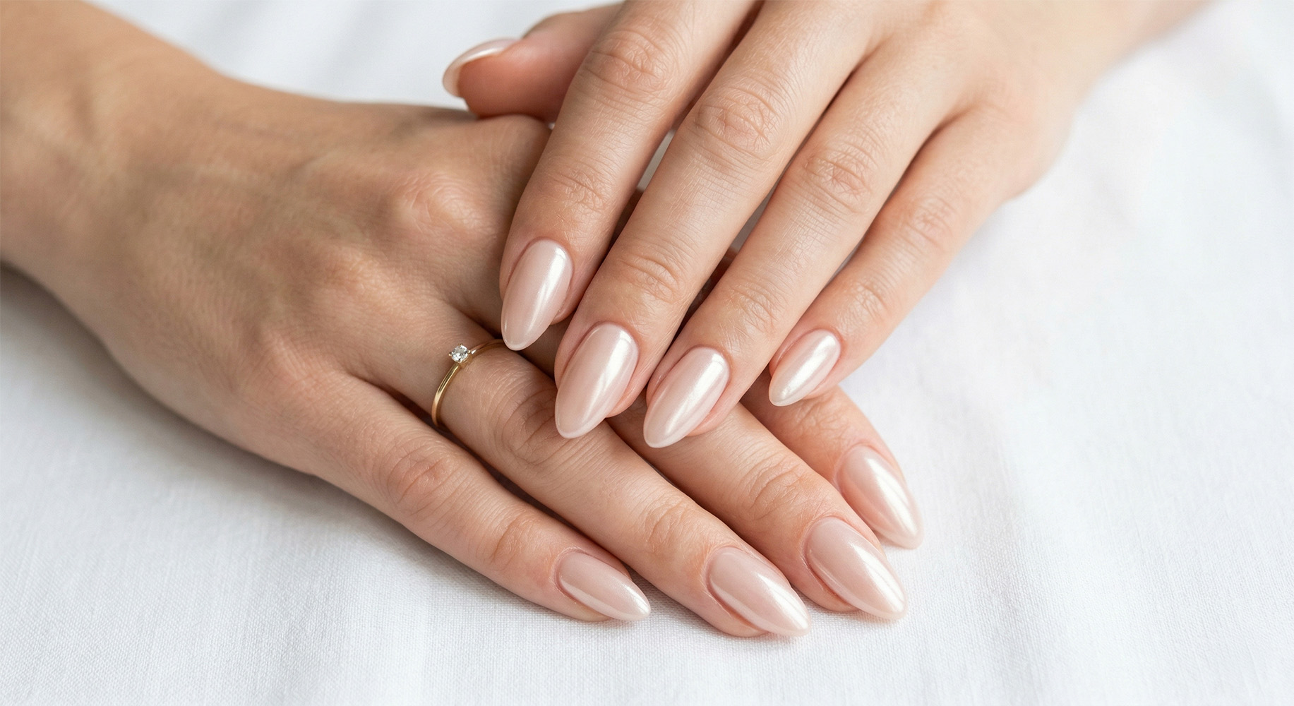 Zbliżenie na eleganckie, migdałowe paznokcie w manicure 'glazed donut nails' lub 'soft chrome', prezentujące mleczno-nude bazę z perłowym chromowym proszkiem, dającą świetlisty blask jak lukier na pączku. Delikatny połysk pięknie chwyta światło. Na palcu złoty pierścionek solitaire