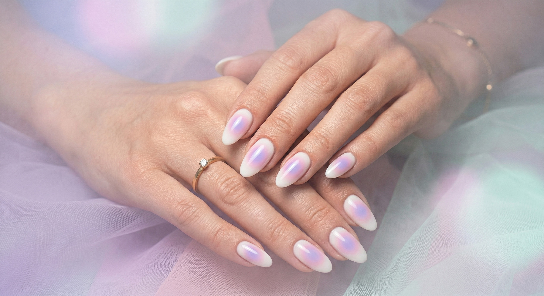 Zadbane kobiece dłonie prezentujące manicure z efektem akwareli i ombre w odcieniach różu, lawendy i błękitu, z delikatnymi zdobieniami kwiatowymi, na tle świeżych białych kwiatów. Na palcu złoty pierścionek z brylantem